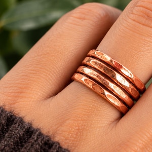 May include: A set of four stacked rings made of polished copper. Each ring features a distinct hammered texture across its surface, creating a series of small, reflective indentations that catch the light. The bands are uniform in width and thickness.
