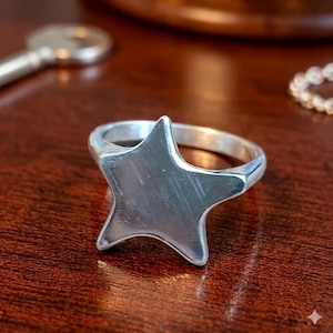 May include: A silver-colored star-shaped ring. The ring features a five-pointed star design with a smooth, polished surface. The ring is set against a dark wooden surface.