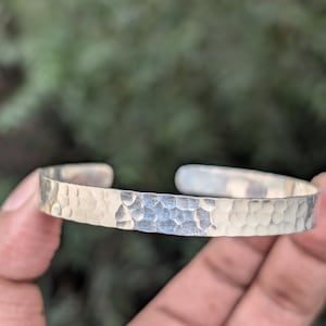 May include: A silver cuff bracelet with a hammered texture. The open-ended bracelet is a light silver colour and has a slightly reflective surface. The bracelet is shown against a blurred green background.