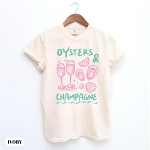 Puede incluir: Camiseta de color marfil con un diseño gráfico en rosa y verde. El diseño incluye las palabras "Oysters & Champagne", ilustraciones de copas de champán, ostras y rodajas de limón. La camiseta está colgada en una percha de madera.