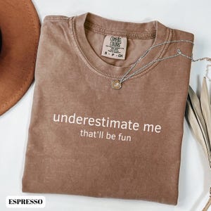 Könnte beinhalten: Espressofarbenes T-Shirt mit dem weißen Text "underestimate me that'll be fun". Das Shirt hat einen Rundhalsausschnitt und ein Comfort Colors-Etikett. Eine silberne Halskette mit einem kleinen Anhänger liegt über dem Shirt.