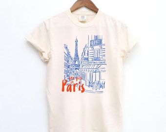 Puede incluir: Camiseta de color marfil con un diseño azul y rojo de una calle parisina, con la Torre Eiffel. El texto "See you in Paris" es rojo. La camiseta está colgada en una percha de madera.