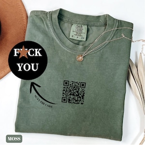 Può includere: T-shirt verde muschio con la scritta "F*CK YOU" in un cerchio nero e un codice QR. La maglietta ha anche la scritta "SCAN THE CODE" con una freccia che punta al codice QR. Una collana d'oro è drappeggiata sulla maglietta.