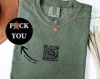 May include: Moss green t-shirt with the text "F*CK YOU" in a black circle, and a QR code. The shirt also has the text "SCAN THE CODE" with an arrow pointing to the QR code. A gold necklace is draped across the shirt.