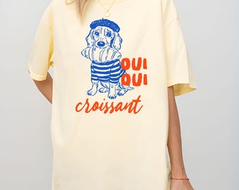 Puede incluir: Camiseta de color plátano con un perro de dibujos animados con boina y camisa a rayas, sosteniendo un cruasán. Las palabras "Oui Oui croissant" están en rojo. La camiseta es de corte holgado.