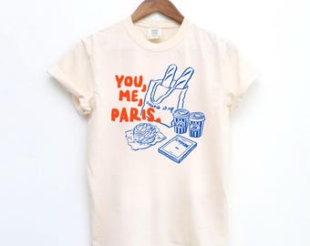 Puede incluir: Camiseta de color marfil con un diseño gráfico. El diseño presenta el texto "YOU, ME, PARIS." e ilustraciones de baguettes, tazas de café, un pastel y un libro. La camiseta está colgada de una percha de madera.