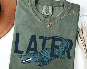 Peut inclure: Un t-shirt vert mousse avec le mot "LATER" en bleu marine, orné d'un motif d'alligator sarcelle. Le t-shirt a un col rond et des manches courtes. Un collier en or avec un pendentif est drapé sur le t-shirt.