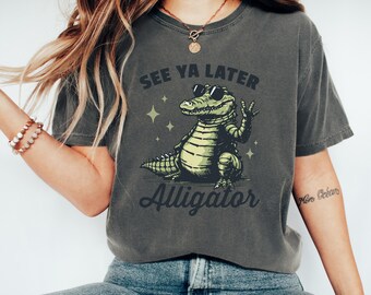 May include: Dark gray t-shirt with the text "SEE YA LATER Alligator" and a cartoon alligator wearing sunglasses. The alligator is green and yellow, with a peace sign hand gesture. The shirt is a relaxed fit.