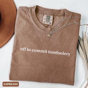 May include: Espresso-coloured t-shirt with the phrase "off to commit tomfoolery" in white. The shirt has a crew neck and short sleeves. A silver necklace with a pendant is draped across the shirt. The shirt is labelled "Comfort Colors".