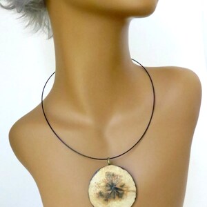 Puede incluir: Un collar con cord&oacute;n negro y un colgante redondo con un dise&ntilde;o de flor prensada. El colgante es de color crema con detalles marrones y negros.