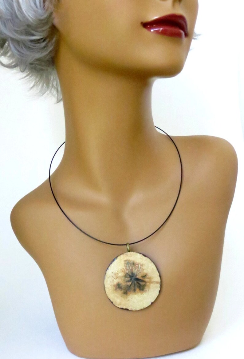 Puede incluir: Un collar con cord&oacute;n negro y un colgante redondo con un dise&ntilde;o de flor prensada. El colgante es de color crema con detalles marrones y negros.