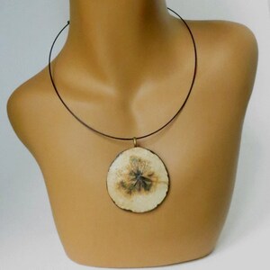 Puede incluir: Un collar con un colgante redondo de color crema y un cord&oacute;n negro. El colgante tiene un dise&ntilde;o floral marr&oacute;n y verde.