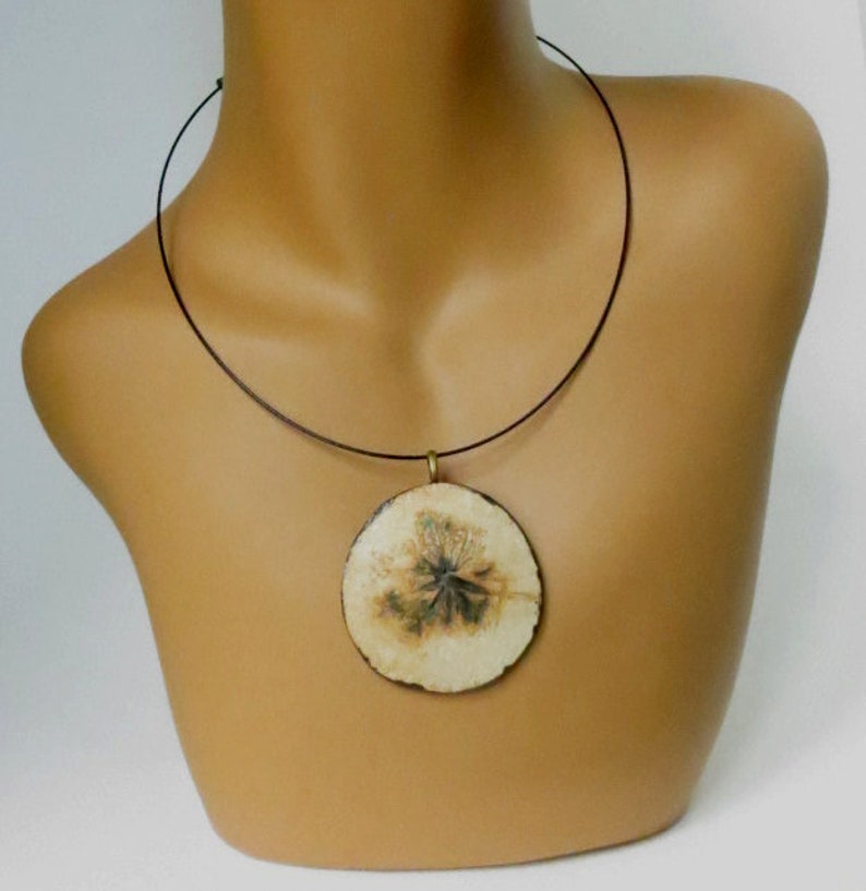 Puede incluir: Un collar con un colgante redondo de color crema y un cord&oacute;n negro. El colgante tiene un dise&ntilde;o floral marr&oacute;n y verde.