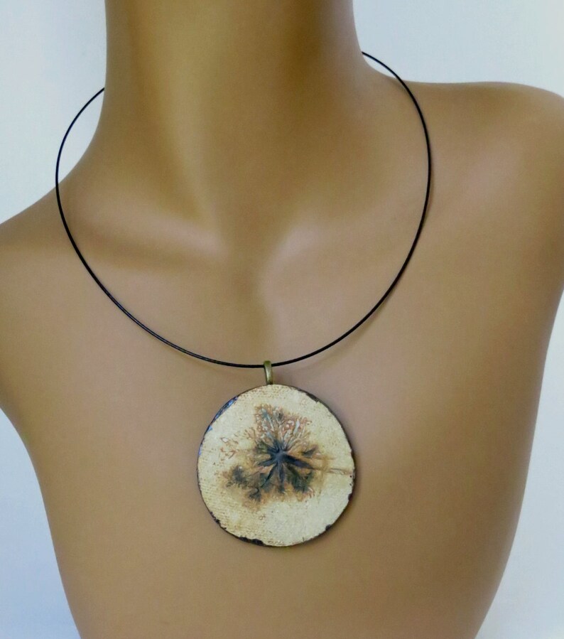 Puede incluir: Un collar con un colgante redondo de cer&aacute;mica blanca con un dise&ntilde;o floral negro. El colgante est&aacute; colgado de un cord&oacute;n negro.