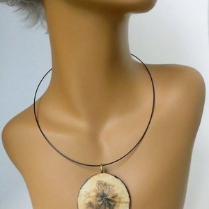 Puede incluir: Un colgante de cer&aacute;mica blanca con un dise&ntilde;o floral marr&oacute;n cuelga de un collar de cord&oacute;n negro. El colgante es redondo y tiene una anilla de metal dorado.