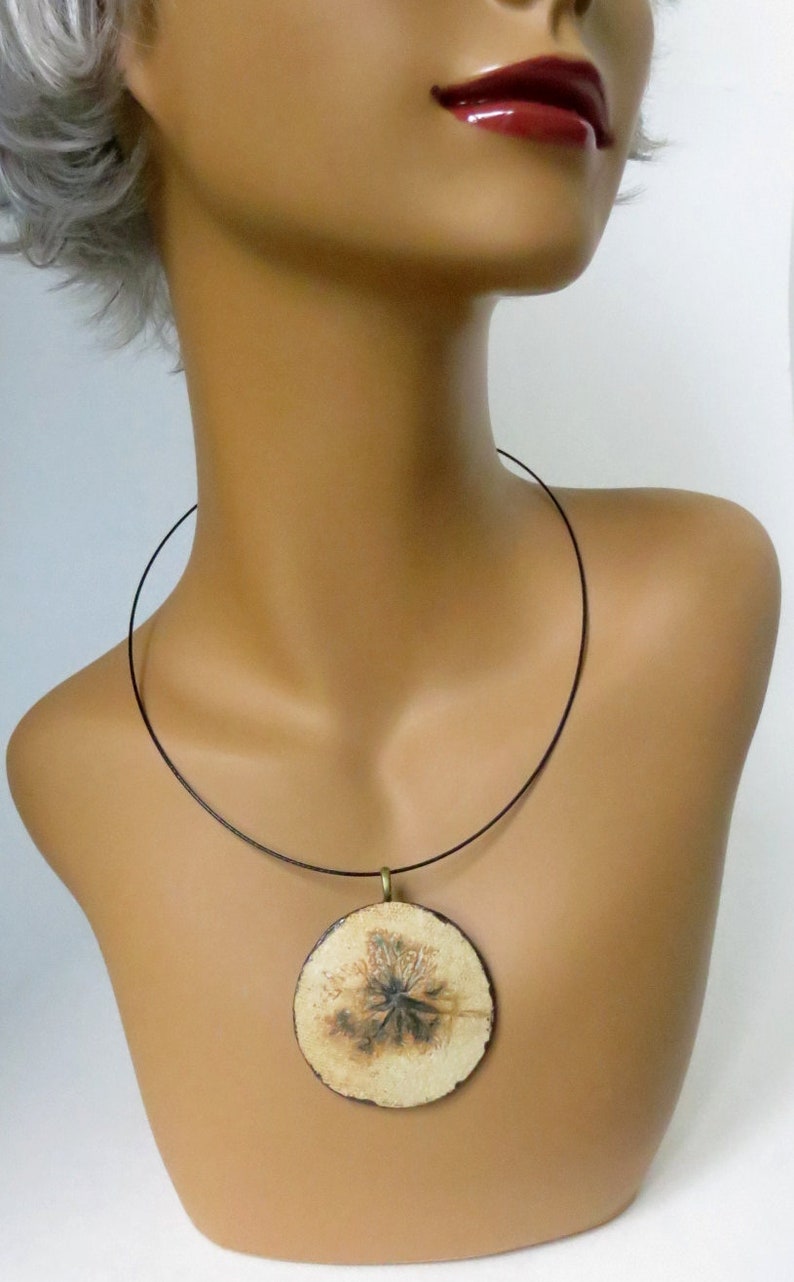 Puede incluir: Un colgante de cer&aacute;mica blanca con un dise&ntilde;o floral marr&oacute;n cuelga de un collar de cord&oacute;n negro. El colgante es redondo y tiene una anilla de metal dorado.