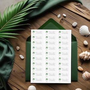 May include: A collection of white address labels with a green palm tree motif, presented within a dark green envelope. Each label displays the text "Rebecca + Henry" and address information. The arrangement is on a wooden surface, with seashells and foliage.