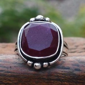 May include: A silver ring with a large, square-cut, deep red gemstone. The gemstone is set in a detailed silver band with small decorative accents. The ring is displayed on a wooden surface, highlighting its design.