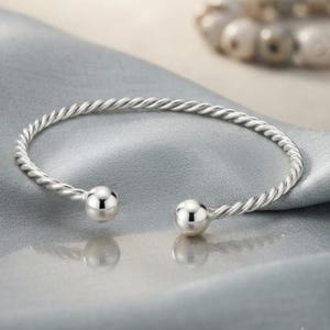 Può includere: Un bracciale a fascia color argento caratterizzato da una banda metallica intrecciata simile a una corda. Il design aperto termina su entrambi i lati con una perla sferica liscia e lucida. La superficie metallica ha una finitura brillante.