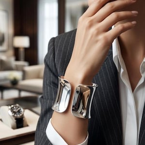 May include: A polished silver cuff bracelet with a curved, open design. The bracelet is worn on a wrist, reflecting light. The person is wearing a gray pinstripe suit and a white collared shirt. A watch is visible in the background.