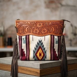 Könnte beinhalten: Rechteckige Handtasche mit gemustertem Stoffkörper und Oberteil aus Leder. Der Stoff hat ein geometrisches Design in Creme, Burgunderrot, Türkis und Gelb. Das Oberteil aus Leder ist mit Blumenschnitzereien und einem Reißverschluss verziert. Braune Fransen hängen an den Seiten.