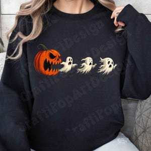 Pode incluir: Camisola preta com um design de Halloween. Uma abóbora laranja com um rosto esculpido está à esquerda, seguida por três fantasmas brancos de desenho animado. O design tem um tema divertido e assustador.