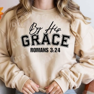 May include: A beige sweatshirt with the text "By His GRACE ROMANS 3:24" in black. The word "GRACE" is in a bold, collegiate-style font. The sweatshirt has long sleeves and a crew neck.