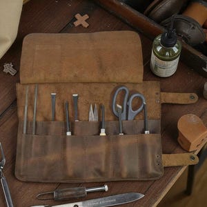 Può includere: Un rotolo di attrezzi in pelle marrone con vari strumenti in metallo, tra cui forbici, scalpelli e lime. Il rotolo è aperto, mostrando gli strumenti in tasche individuali. Una piccola bottiglia e altri strumenti sono visibili.