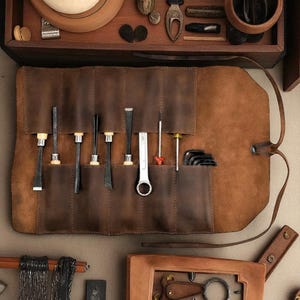 May include: A brown leather tool roll with various tools, including chisels, a wrench, and Allen wrenches. The roll is open, displaying the tools in individual pockets. The leather has a natural, textured appearance, and the tools are made of metal.