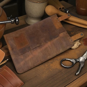 May include: A brown leather tool roll with multiple slots, secured by two straps. The roll is open, revealing the interior. The leather has a distressed look. Other tools and accessories are scattered around the roll on a wooden surface.