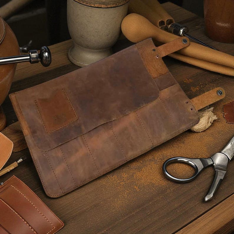May include: A brown leather tool roll with multiple slots, secured by two straps. The roll is open, revealing the interior. The leather has a distressed look. Other tools and accessories are scattered around the roll on a wooden surface.