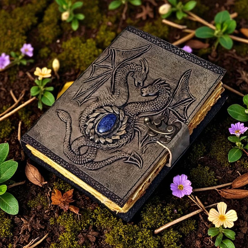 Handcrafted fantasy leather journal with embossed twin dragons, gemstone centerpiece, and brass lock, resting on mossy ground with wildflowers.