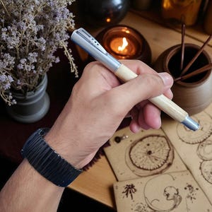 Puede incluir: Un bol&iacute;grafo plateado y de madera con un cuerpo de madera clara y detalles plateados se sostiene en una mano. Una pulsera negra est&aacute; en la mu&ntilde;eca. El bol&iacute;grafo est&aacute; cerca de un jarr&oacute;n con flores secas, una vela encendida y papeler&iacute;a.