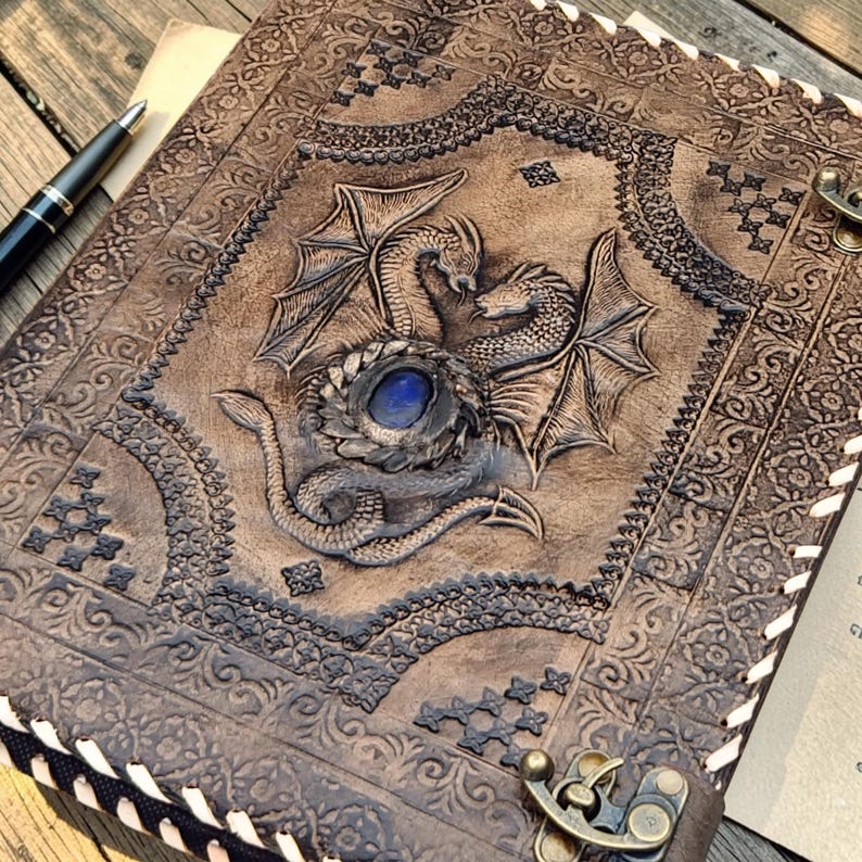 A4 leather ring binder journal with dragon embossing and blue gemstone detail, styled on parchment fabric and wooden background