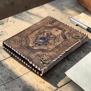 Leather dragon embossed A4 ring binder journal with blue gemstone accent, placed on rustic wooden table with parchment fabric, cinematic warm lighting and high detail fantasy stationery