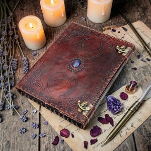Puede incluir: Un diario de cuero marrón oscuro con diseños de luna creciente en relieve y una gema azul central. El diario tiene cierres de latón y está rodeado de velas, lavanda seca e instrumentos de escritura.