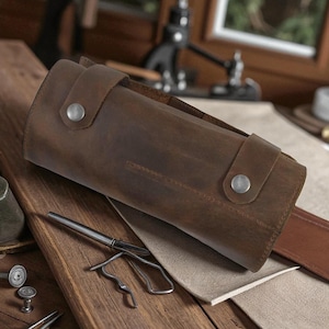 May include: A brown leather tool roll with silver snap closures. The roll is cylindrical and appears to be made of distressed leather. A metal tool and other items are visible on a wooden surface.