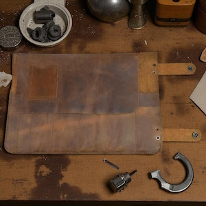 May include: A brown leather tool roll, open and laid flat, with multiple slots for tools. The roll has a flap closure with a snap. Several tools and parts are scattered around the roll on a wooden surface. The leather has a distressed appearance.
