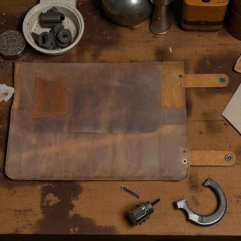 May include: A brown leather tool roll, open and laid flat, with multiple slots for tools. The roll has a flap closure with a snap. Several tools and parts are scattered around the roll on a wooden surface. The leather has a distressed appearance.