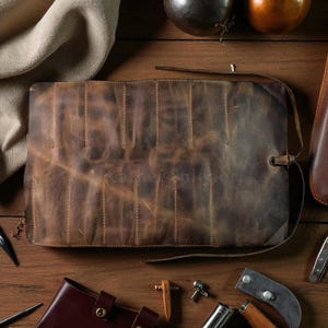 May include: A brown leather tool roll with multiple slots, secured with a leather strap. The roll is displayed on a wooden surface, surrounded by various tools and accessories. The leather has a distressed finish.