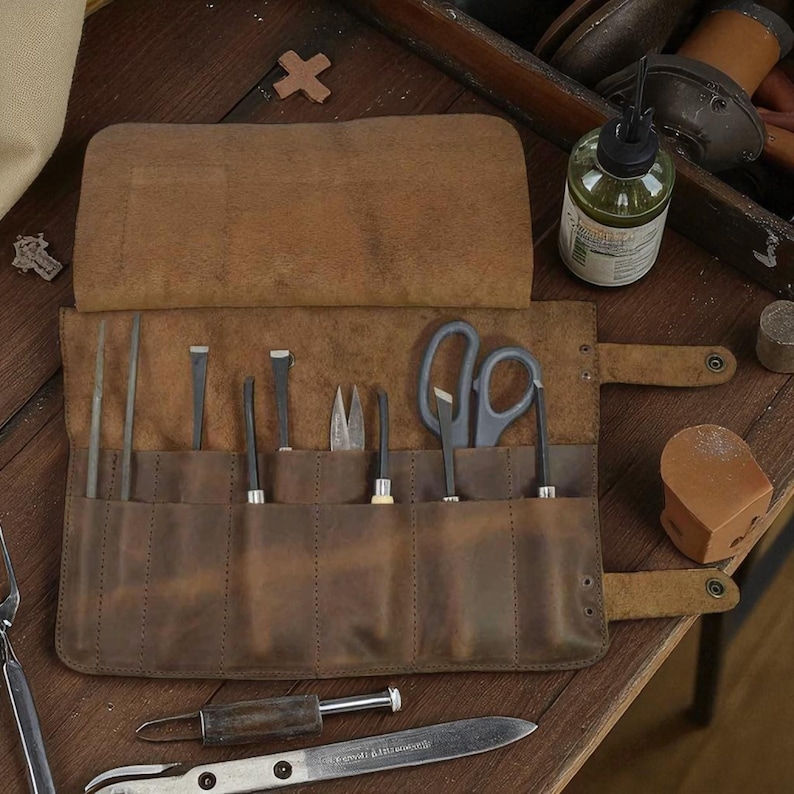 May include: A brown leather tool roll, open to display various metal tools, including scissors, chisels, and files. The roll is on a wooden surface, with a bottle of oil and other tools visible in the background. The roll is designed to hold and organize tools.