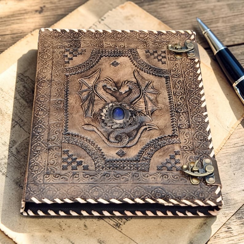 Leather dragon embossed A4 ring binder journal with blue gemstone centerpiece on rustic wooden desk, fantasy antique stationery