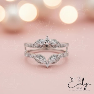 Peut inclure: Une bague de protection en argent avec un motif floral, ornée de pierres précieuses claires de taille marquise et ronde. La bague a une tige fendue et est conçue pour être portée avec une autre bague. Le fond est rose clair.