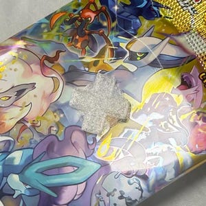 May include: A clear, cross-shaped object rests on a colorful, illustrated surface. The background features various cartoon characters in shades of blue, purple, yellow, and white. The cross appears to be made of a sparkly, translucent material.
