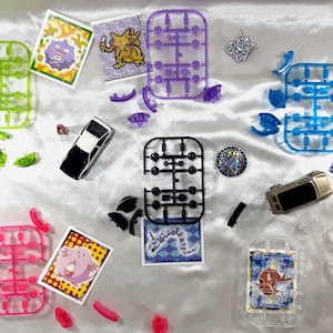 Könnte beinhalten: Eine Ansammlung farbenfroher Kunststoff-Modellbausatzteile, darunter grüne, lila, blaue, rosa und transparente Rahmen, zusammen mit kleinen Spielzeugautos und Pokémon-Sammelkarten, angeordnet auf einer weißen Oberfläche.