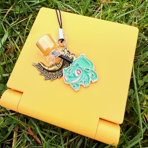 Op de afbeelding: Een gele telefoonhoes met een zwart koord en zilveren sluiting. Bevestigd zijn een drankbedel, een bronzen blad en een groene Bulbasaur-bedel. De items liggen op groen gras.