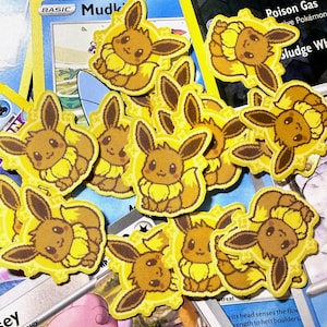 Peut inclure: Plusieurs autocollants jaunes et bruns représentant le personnage Eevee sont disposés sur des cartes à collectionner Pokémon. Les autocollants présentent le personnage Eevee avec ses oreilles et sa queue touffue. Les cartes ont du texte et des illustrations.