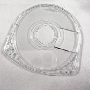 May include: A clear, transparent plastic tape dispenser with a circular design. The dispenser has a central opening and a cutting edge for the tape. The design includes rounded edges and a smooth surface, suitable for office or craft use.
