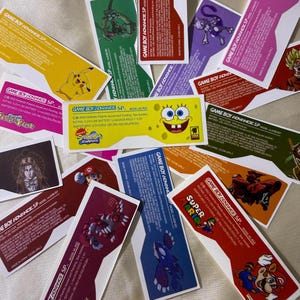May include: A selection of Game Boy Advance SP labels with diverse designs and characters. The labels are rectangular with vibrant backgrounds, showcasing characters like Pikachu, SpongeBob, and Mario. Text is present on the labels.
