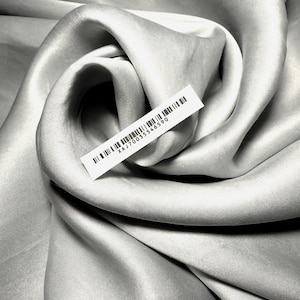 Peut inclure: Gros plan sur un tissu gris clair avec des plis doux et un léger éclat. Une étiquette blanche avec un code-barres et le texte "XAJ70035546590" est attachée. Le tissu semble luxueux, peut-être de la soie ou du satin.